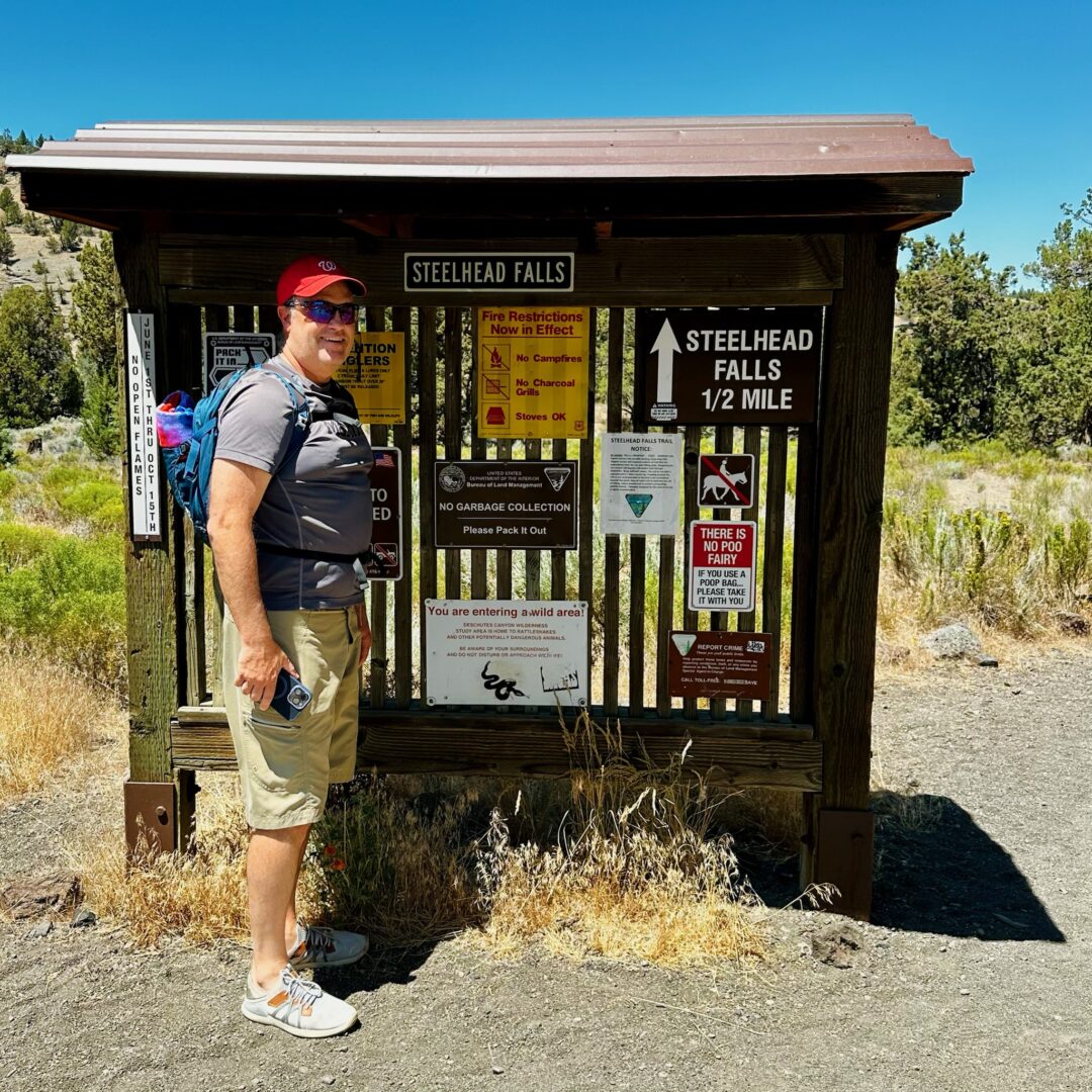 A person in a red cap, sunglasses, blue shirt, beige shorts, and sneakers stands smiling beside a Steelhead Falls trail signboard filled with notices and maps. They carry a backpack amid sunny surroundings of dry grass and sparse trees.