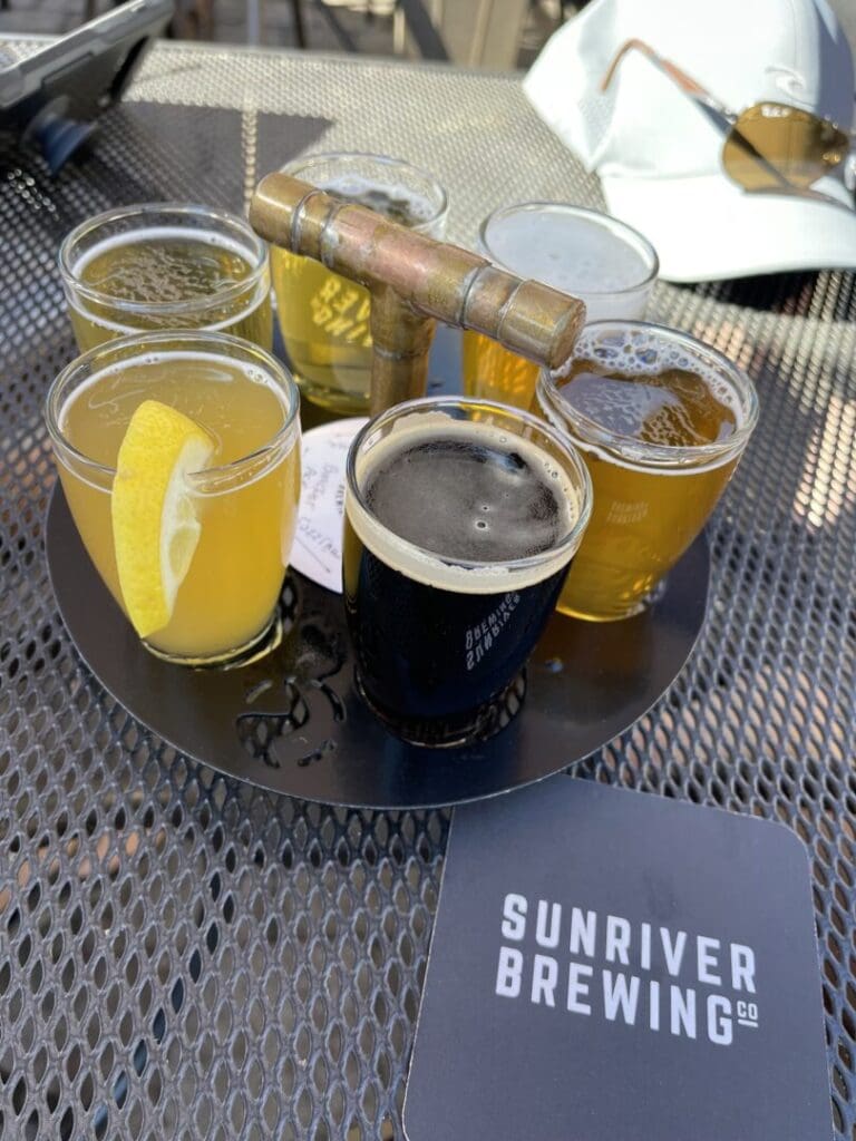 a Round Metal Tray on an Outdoor Metal Table Holds Six Small Glasses of Beer One with a Lemon Slice the Tray Has a Brass Handle a Sunriver Brewing Co Coaster and Sunglasses Hint at the Great Breweries in Bend Oregon