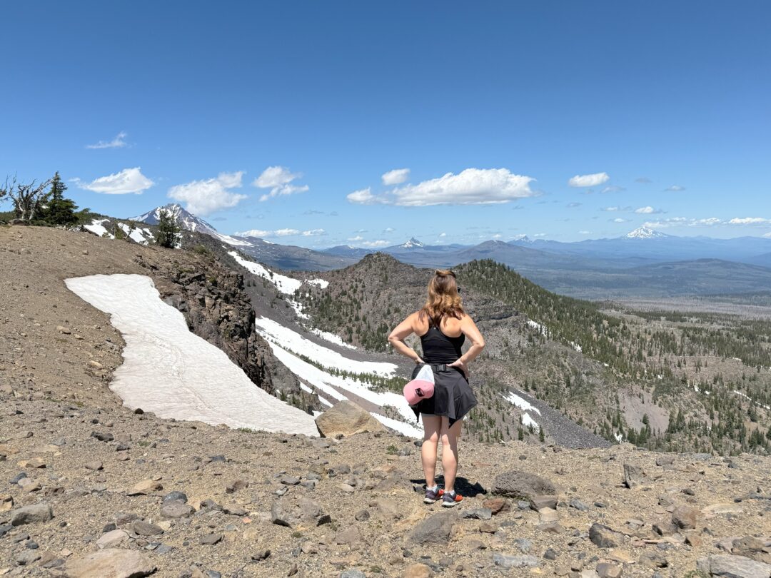 Tam Mcarthur Rim Trail Hike Summit View a Woman Stands on a Rocky Mountain Trail Near Steelhead Falls Looking at Distant Snow Capped Peaks and Green Forests Below She Wears a Black Tank Top Shorts Sneakers and Carries a Pink Rolled Up Mat Patches of Snow Dot the Ground Beneath a Clear Blue Sky