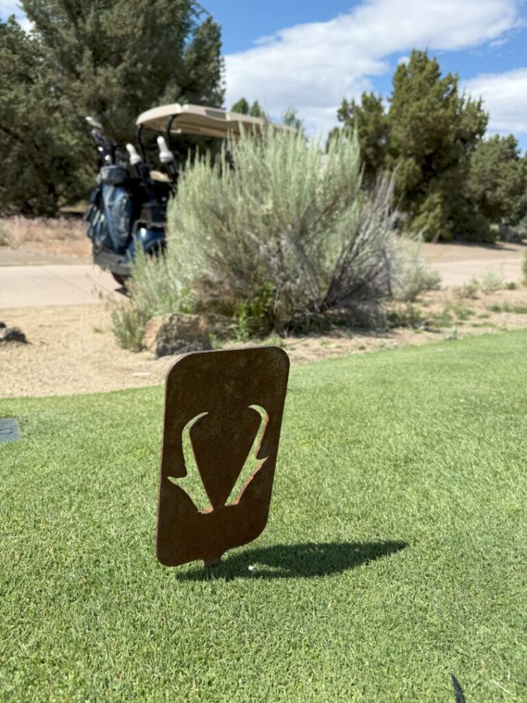 Tee Marker Pronghorn Golf Club Bend a Metal Tee Marker with an Antelope Horn Design Stands on a Green Pronghorn Golf Course Tee Box in the Background Are Golf Bags on a Cart Bushes Rocks and Trees Beneath a Partly Cloudy Sky Highlighting This Scenic Outdoor Golf Experience