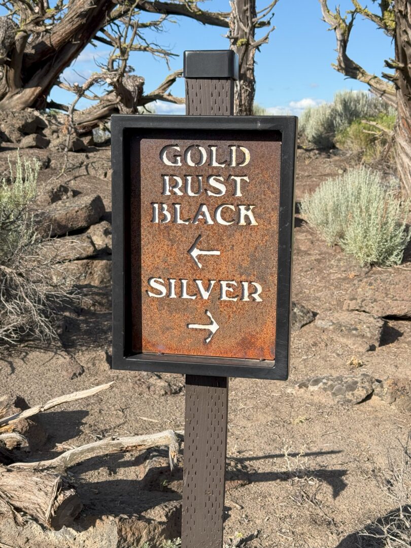 Tee Markers Pronghorn Golf a Weathered Metal Sign on a Post Reads Gold Rust Black with a Left Arrow and Silver with a Right Arrow Set Outdoors Near Pronghorn Golf Course It Stands in a Dry Rocky Area Dotted with Sparse Shrubs and Twisted Tree Branches