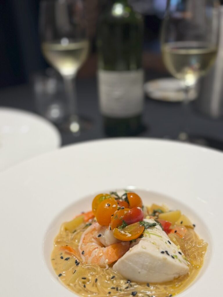 Thai curry market table bistro a Close up of a Plated Gourmet Dish from Market Table Bistro Featuring White Fish Shrimp Colorful Cherry Tomatoes and Sesame Seeds over Noodles in a Creamy Sauce in the Blurred Background Two Glasses of White Wine and a Wine Bottle Are Visible on a Dark Table