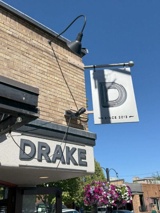 A beige brick building with a black sign reading “DRAKE” above the entrance, one of the standout Bend Restaurants. A white hanging sign with a large “D” and “SINCE 2013” is above it, surrounded by flowers, street lamps, and a clear blue sky.