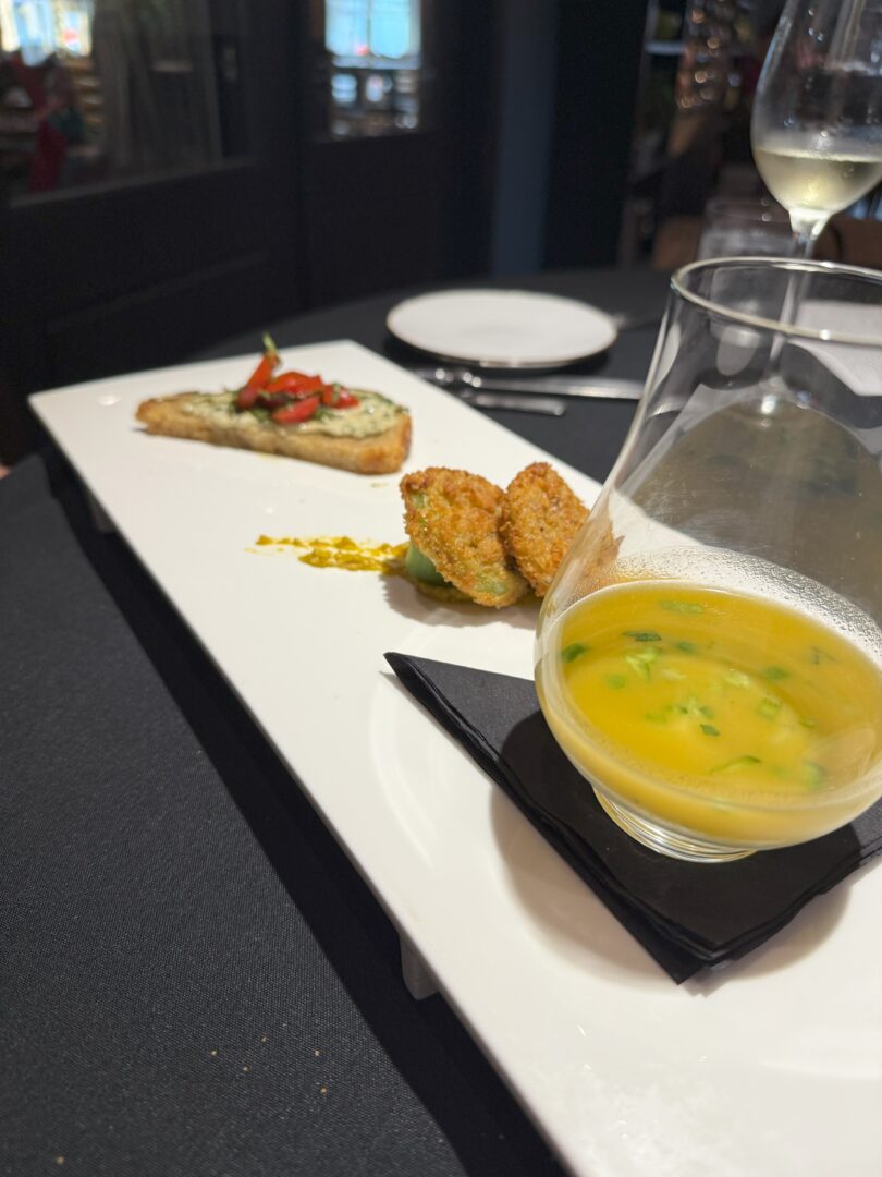 Tomato starter market table bistro at Market Table Bistro a White Rectangular Plate Holds a Glass of Yellow Soup with Herbs Two Breaded Croquettes and a Slice of Toasted Bread Topped with Diced Vegetables the Plate Rests on a Dark Tablecloth Wine Glasses Blurred in the Background