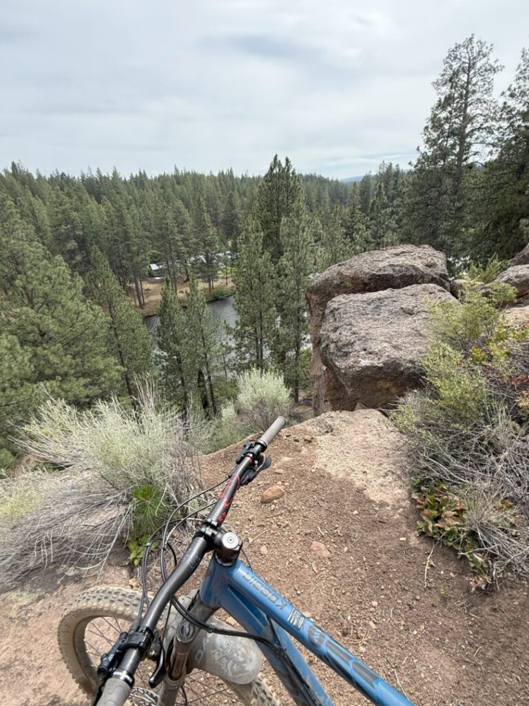 A mountain bike rests on Rimrock Trail at the edge of a rocky cliff, overlooking a forest of tall pine trees and a small river below. The sky is overcast, with bushes and shrubs lining the trail in the foreground.