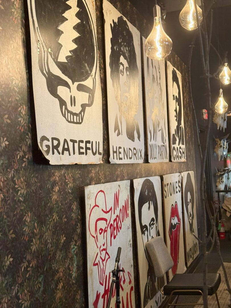 A wall with six large, stylized black-and-white portraits of famous musicians, including Grateful Dead, Hendrix, Dolly Parton, Johnny Cash, Hank Williams, and the Rolling Stones. Exposed bulbs hang from the ceiling, and a chair and mic stand sit below the paintings.