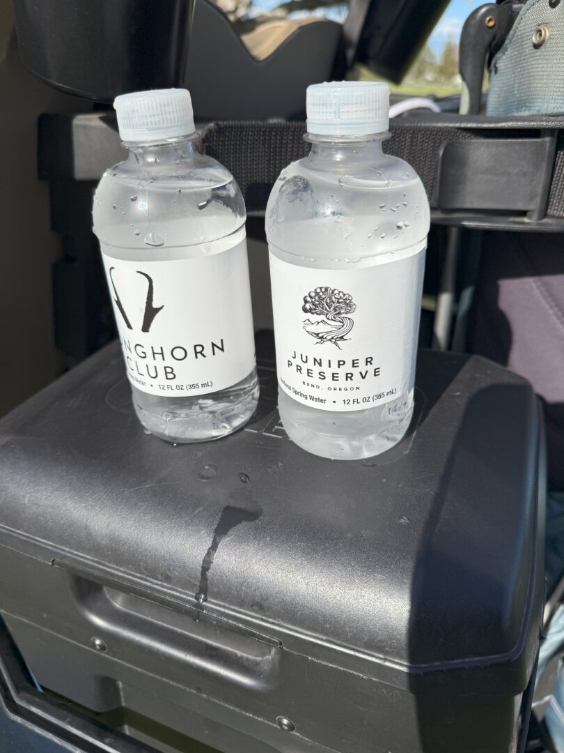 Water Bottles Pronghorn Golf Club Two Clear Plastic Water Bottles with White Labels Sit on a Black Surface the Left Label Reads Longhorn Club with a Horn Logo Perfect for Pronghorn Golf Course the Right Juniper Preserve with a Juniper Tree Logo Both Are Half Full and Have Condensation