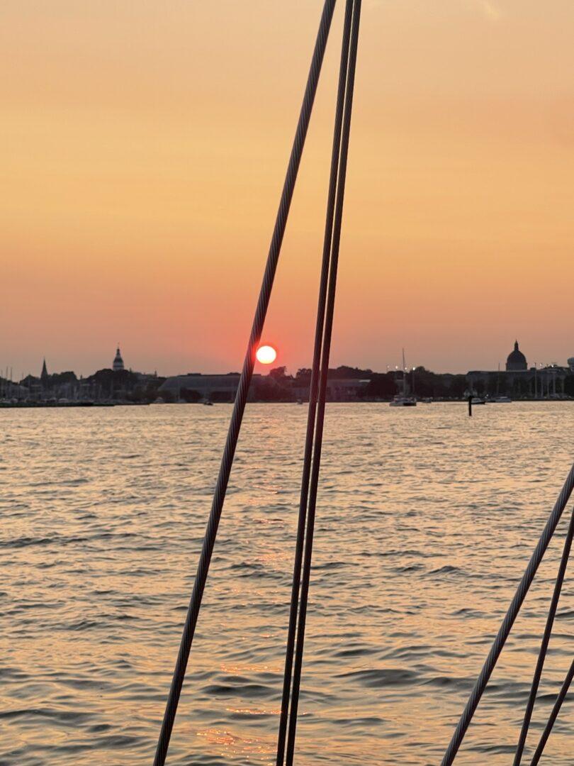 Sunset Annapolis Md Harbor a Vivid Sunset over Calm Water in Annapolis Maryland with the Orange Sun Low in the Sky Sailboat Rigging Lines Cross the Foreground While Silhouetted Buildings and Boats Line the Distant Shore Under a Soft Orange and Yellow Sky