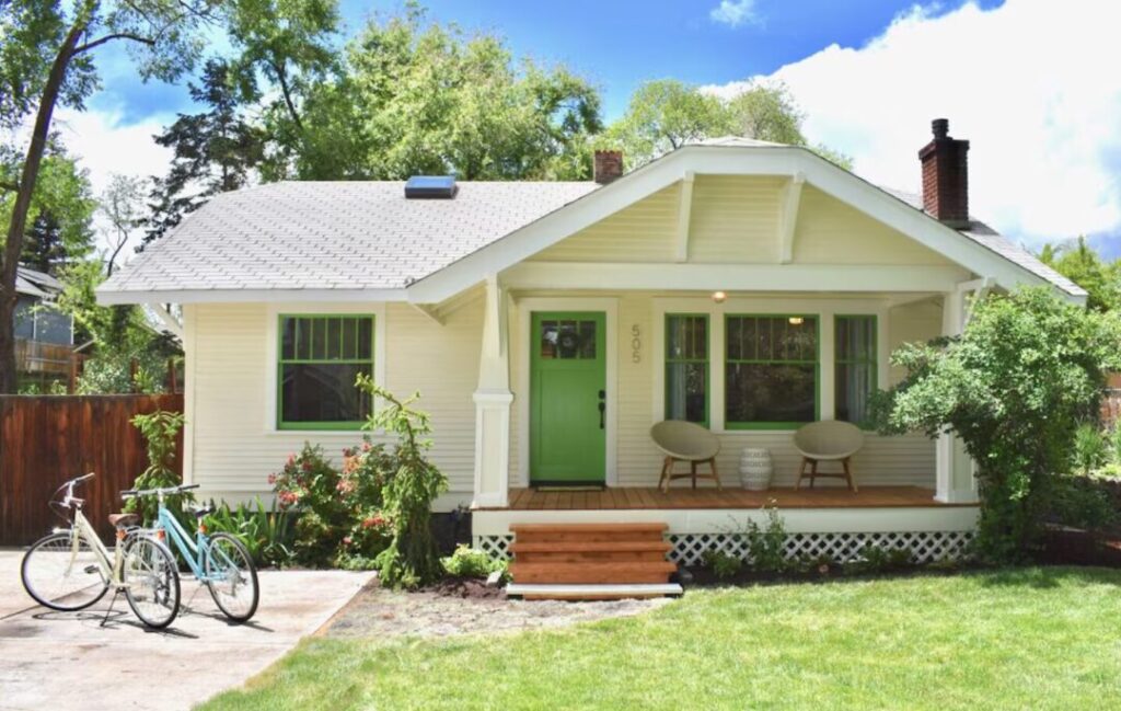 Airbnb in Bend Oregon a Charming Single Story Cream House with a Green Door and Matching Window Frames in Bend Oregon Perfect for an Airbnb Stay It Features a Cozy Porch Two Bikes and is Surrounded by a Lush Lawn Under the Bright Blue Sky