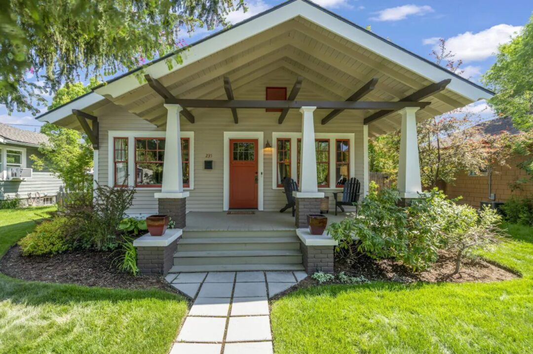 Downtown-bend-oregon-bungalow A charming craftsman-style house—an ideal option if you’re wondering where to stay in Bend, Oregon. Enjoy the front porch with white columns, a red door, brown trim, and lush greenery all around under a blue sky dotted with white clouds.
