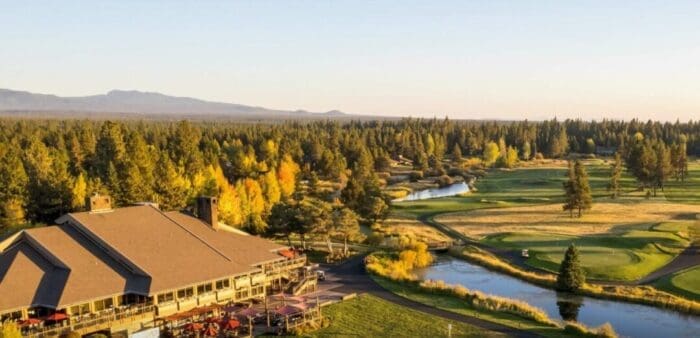 The Sunriver Lodge at Sunriver Resort with its golf course is surrounded by lush green fairways and autumn-colored trees—a picturesque choice for those seeking where to stay in Bend, Oregon, with mountain views under a golden sunset sky.