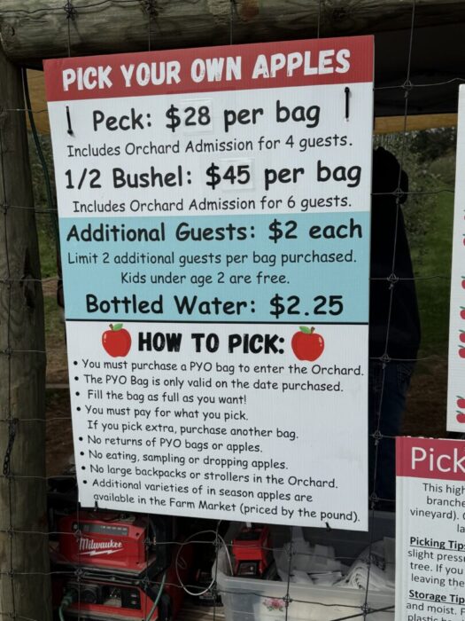 A sign at Gaver Farm titled “Pick Your Own Apples” lists prices: 1 Peck is $28 per bag, 1/2 Bushel is $45 per bag—both include orchard admission for 6 guests. Extra guests are $2 each and bottled water is $2.25. Apple-picking instructions follow below.