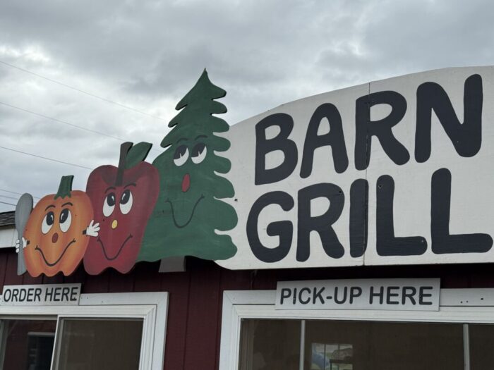 A sign for “Barn Grill” at Gaver Farm features cartoon faces on a pumpkin, an apple, and a pine tree above two windows labeled ORDER HERE and PICK-UP HERE. The sky is cloudy and the red building has white trim.