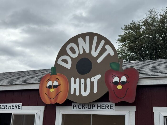 A round sign reading DONUT HUT with a donut hole in the center is displayed at Gaver Farm. Beside the sign are cartoon faces of a smiling pumpkin and apple. The sky is cloudy, with “ORDER HERE” and “PICK-UP HERE” signs below.