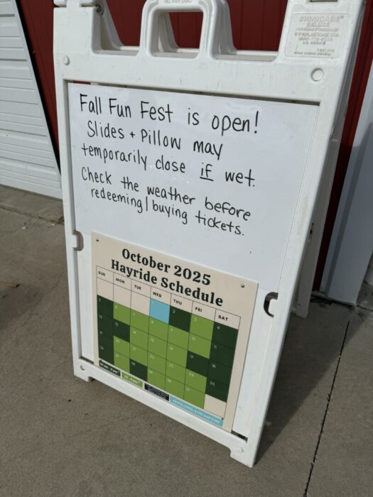 A white sandwich board sign stands on the Gaver Farm sidewalk. The top has handwritten text about Fall Fun Fest, noting slides and pillow may close if wet. Below is an October 2025 hayride schedule calendar in green shades.