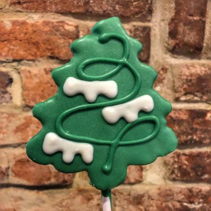 A green Christmas tree-shaped cookie pop from Gaver Farm, decorated with white and green icing garlands and snow-like details, is held in front of a textured brick wall.
