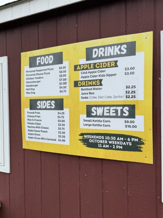 A yellow and white menu board on a red wooden wall at Gaver Farm displays prices for food, drinks, sides, and sweets. Items include pizza, chicken tenders, cheeseburgers, fries, apple cider, popcorn, and more, plus weekend and weekday hours.