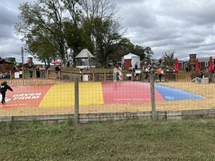 A large colorful jumping pillow with red, yellow, and blue sections sits in a sandy area at Gaver Farm, surrounded by a wooden fence. Children play on the pillow as adults watch nearby among trees, picnic tables, farm buildings, and cloudy skies.