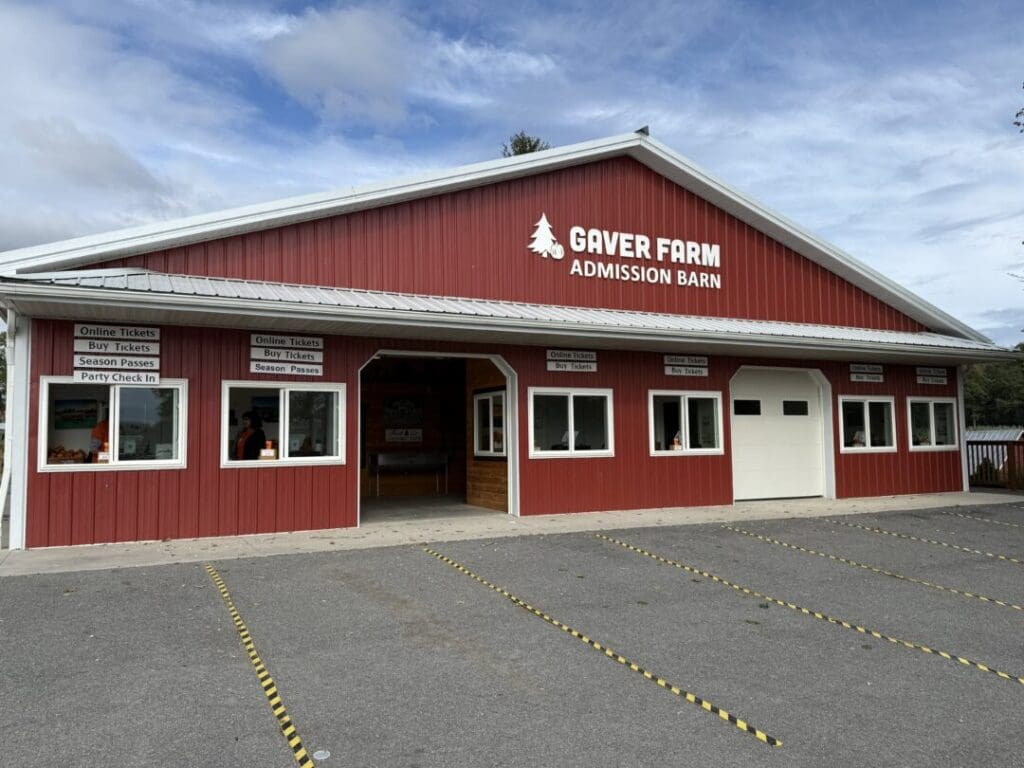 a Red Barn Style Building with White Trim Labeled Gaver Farm Admission Barn Stands Under a Partly Cloudy Sky in Mt Airy Md Signs Above the Windows Mention Buying Tickets and Season Passes While Yellow Lines Mark Parking Spaces in the Foreground