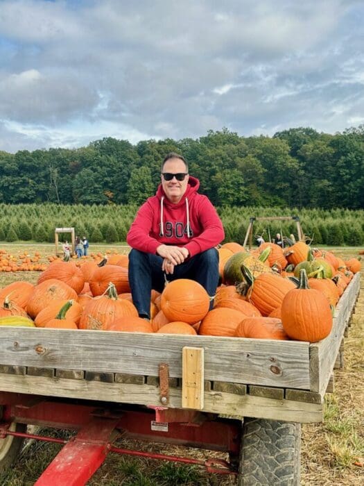 a Man Wearing Sunglasses a Red Hoodie Jeans and Sneakers Sits Cross Legged on a Wooden Wagon Filled with Orange Pumpkins in a Pumpkin Patch in Frederick Mdone of the Fun Things to Do in Frederick on a Cloudy Day