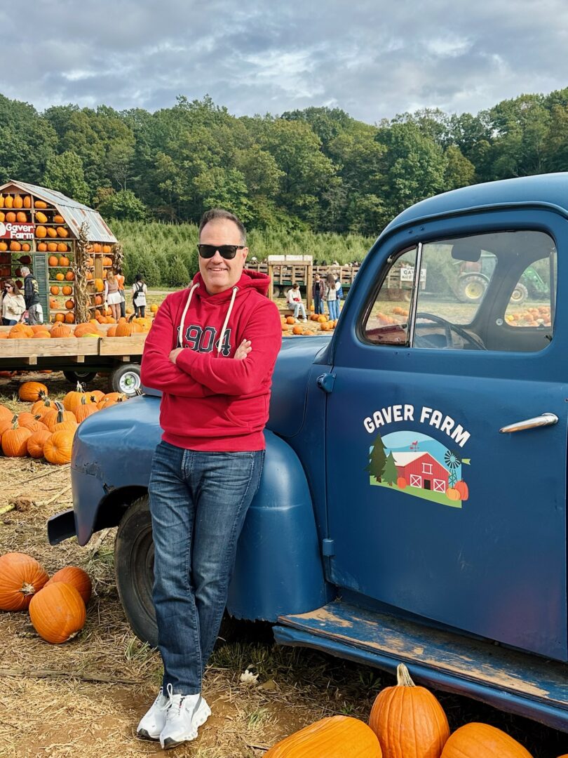 a Man in a Red Hoodie and Jeans Smiles with Arms Crossed Leaning Against a Blue Gaver Farm Vintage Truck Hes Surrounded by Scattered Pumpkins in the Patch Autumn Trees and a Festive Wooden Stand Filled with Pumpkins and Decorations in the Background