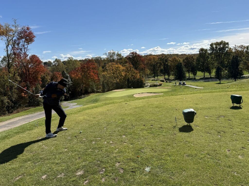 Links at Challedon Elevated Tee Shot a Golfer is Mid swing on a Tee Box at the Links at Challedon Surrounded by Green Grass and Autumn Trees with Red Yellow and Orange Leaves Under a Clear Blue Sky Two Green Baskets Are Nearby with Distant Golf Carts in the Background