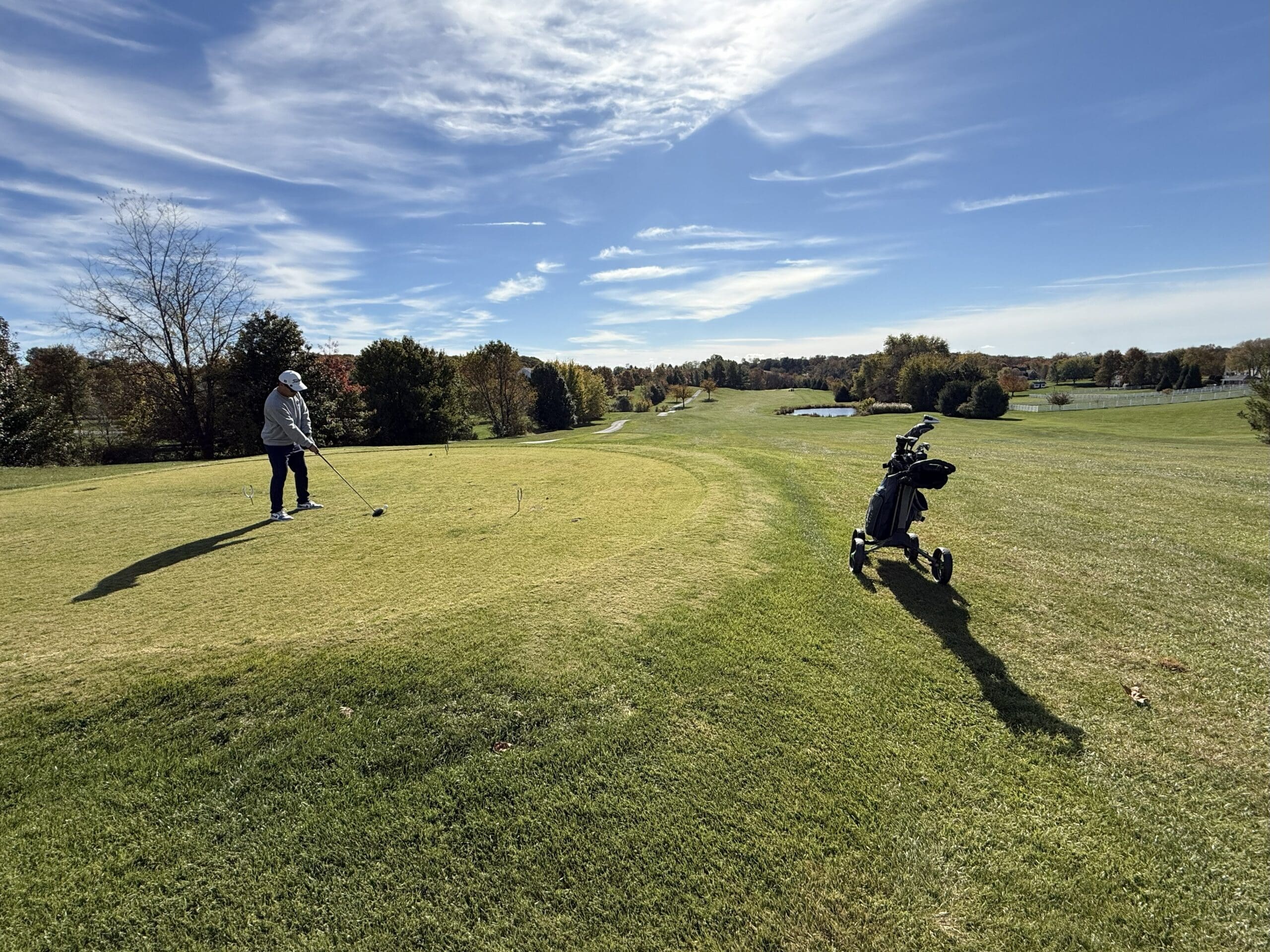 Links at Challedon Hole Two a Golfer Stands on the Tee at the Links at Challedon Preparing to Swing on a Sunlit Course with Autumn Trees in the Distance a Pushcart with Clubs Casts a Long Shadow Across the Grass Under a Partly Cloudy Blue Sky