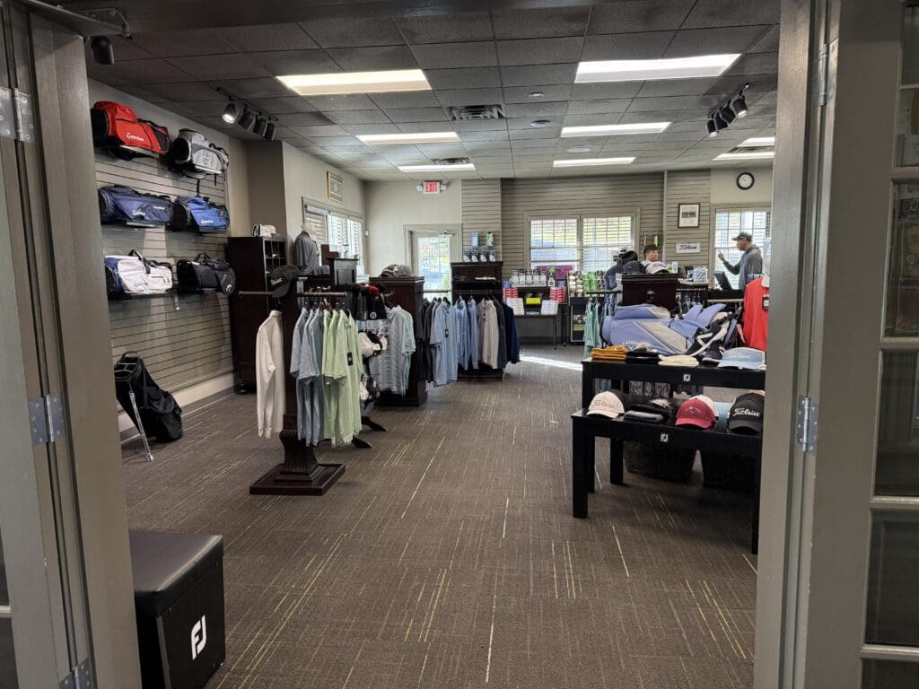 Links at Challedon Pro Shop a Golf Pro Shop Interior at the Links at Challedon with Racks of Clothing Hats and Golf Bags on Display Two People Stand at the Checkout Counter Near the Back the Shop Features Carpeted Floors Bright Lighting and Large Windows Throughout
