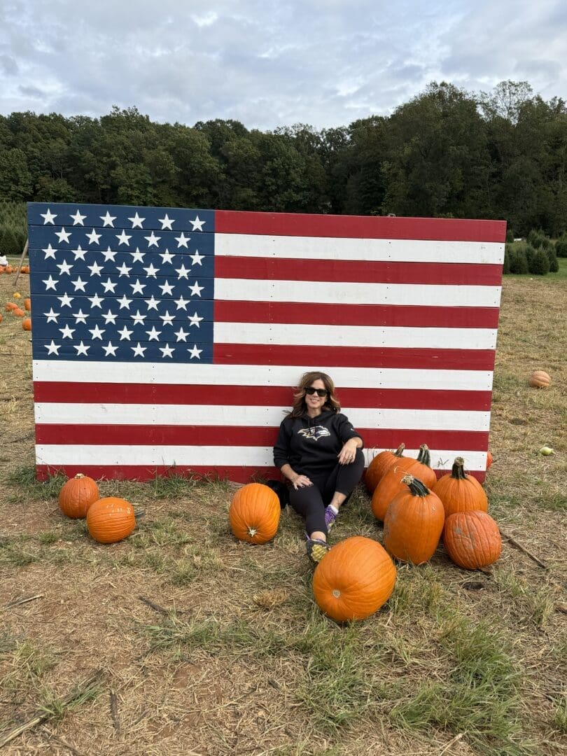 a Woman Wearing Sunglasses Sits on the Ground at Gaver Farm Leaning Against a Large Wooden American Flag Display She is Surrounded by Several Pumpkins on Dry Grass in a Pumpkin Patch with Trees and a Cloudy Sky in the Background