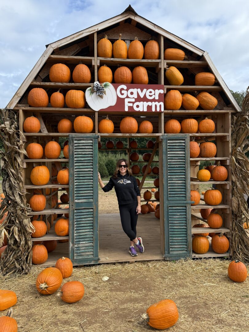 Nicole barn house pumpking patch gaver farm a Woman in Sunglasses and Black Clothes Stands in the Doorway of a Wooden Pumpkin House at Gaver Farm Surrounded by Pumpkins on Shelves and Festive Corn Stalks Hay Covers the Ground As a Gaver Farm Sign Welcomes Visitors to This Autumn Scene