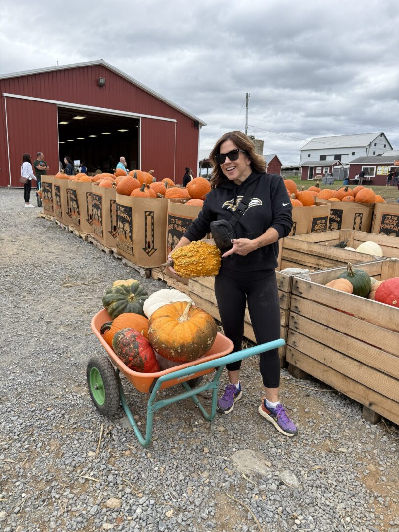 Nicole gaver farm pumpkin picking a Woman in Sunglasses and Athletic Wear Smiles While Holding a Large Yellow Gourd Beside a Wheelbarrow Full of Pumpkins at Gaver Farms Outdoor Pumpkin Patch Behind Her Are Crates of Pumpkins and a Red Barn Under a Cloudy Sky