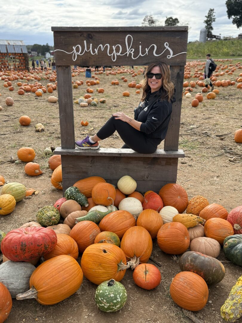 Nicole pumpkin patch gaver farm a Woman with Wavy Brown Hair Wearing Sunglasses a Black Outfit and Purple Shoes Sits Smiling in a Wooden Frame Labeled Pumpkins at Gaver Farm She is Surrounded by Orange and Multicolored Pumpkins at a Lively Pumpkin Patch