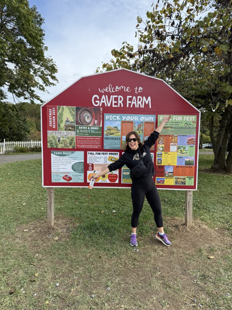 Nicole welcome to gaver farms a Woman in Sunglasses Black Clothes and Purple Sneakers Stands Smiling and Pointing at a Large Red welcome to Gaver Farm Sign Filled with Colorful Information and Farm Rules She Holds a Gaver Farm Flier Standing on Grass Near Trees and a White Fence