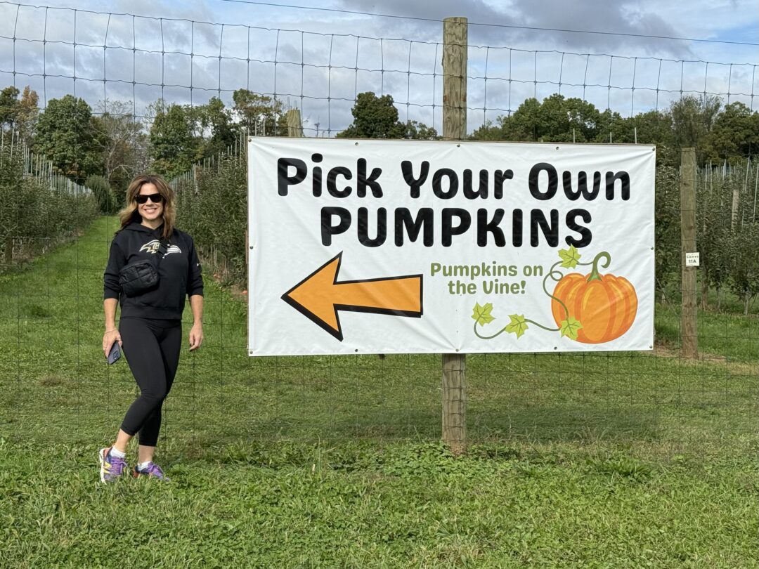 Pyo pumpkins gaver farms md a Woman in Sunglasses and Athletic Wear Stands Smiling Beside a Large pick Your Own Pumpkins Sign at Gaver Farm on a Grassy Field Pumpkin and Vine Illustrations Decorate the Sign Trees Line the Background Under a Cloudy Sky