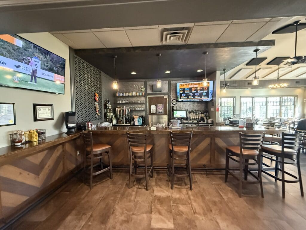the Grille Links at Challedon a Modern Bar at the Links at Challedon Features a Wooden Counter and Four High Chairs Shelves with Bottles and Glasses Line the Back Two Digital Menu Screens Glow and a Tv on the Left Wall Shows Golf Large Windows on the Right Fill It with Light