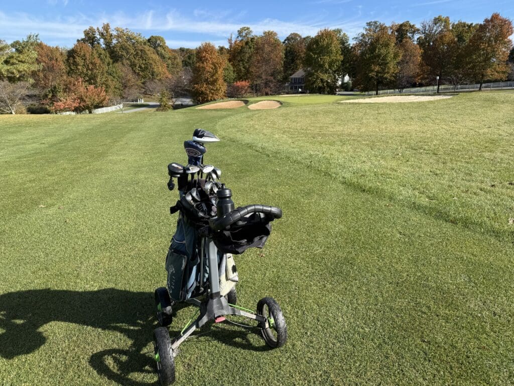 the Links at Challedon Golf Approach Shot a Golf Pushcart Holding Several Clubs Sits on a Lush Green Fairway at the Links at Challedon in the Distance Sand Bunkers Autumn colored Trees and a House Appear Under a Blue Sky with Wispy Clouds and Gentle Morning Shadows