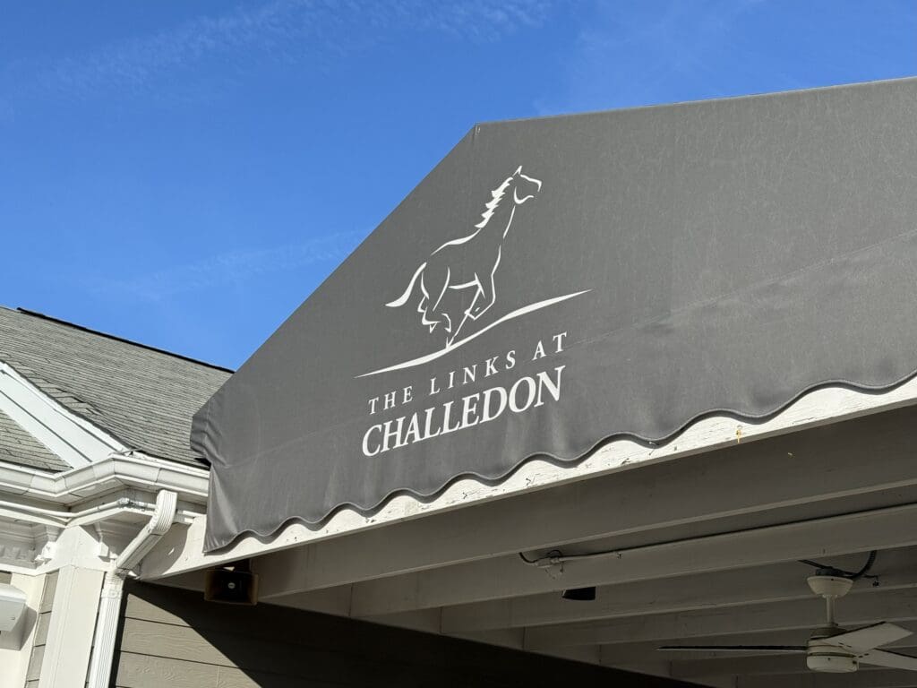 the Links at Challedon Golf Course a Gray Awning Displays a White Outline of a Running Horse Above the Words the Links at Challedon the Awning Covers Part of a Building with White Trim Highlighting the Distinctive Entrance to the Links at Challedon Under a Clear Blue Sky