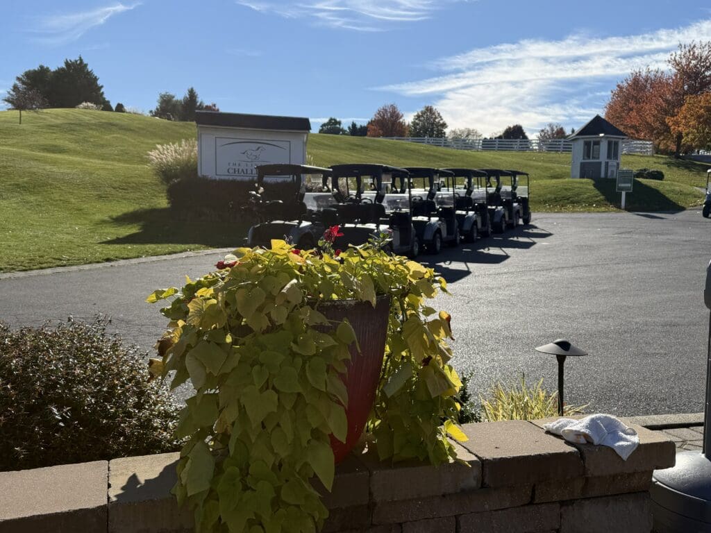 the Links at Challedon Golf Maryland a Row of Golf Carts is Parked on a Paved Lot at the Links at Challedon Near a Grassy Hill with a White Sign and Small Building a Large Planter with Green and Yellow Plants Sits in the Foreground Beneath Blue Wispy Skies and Autumn Trees