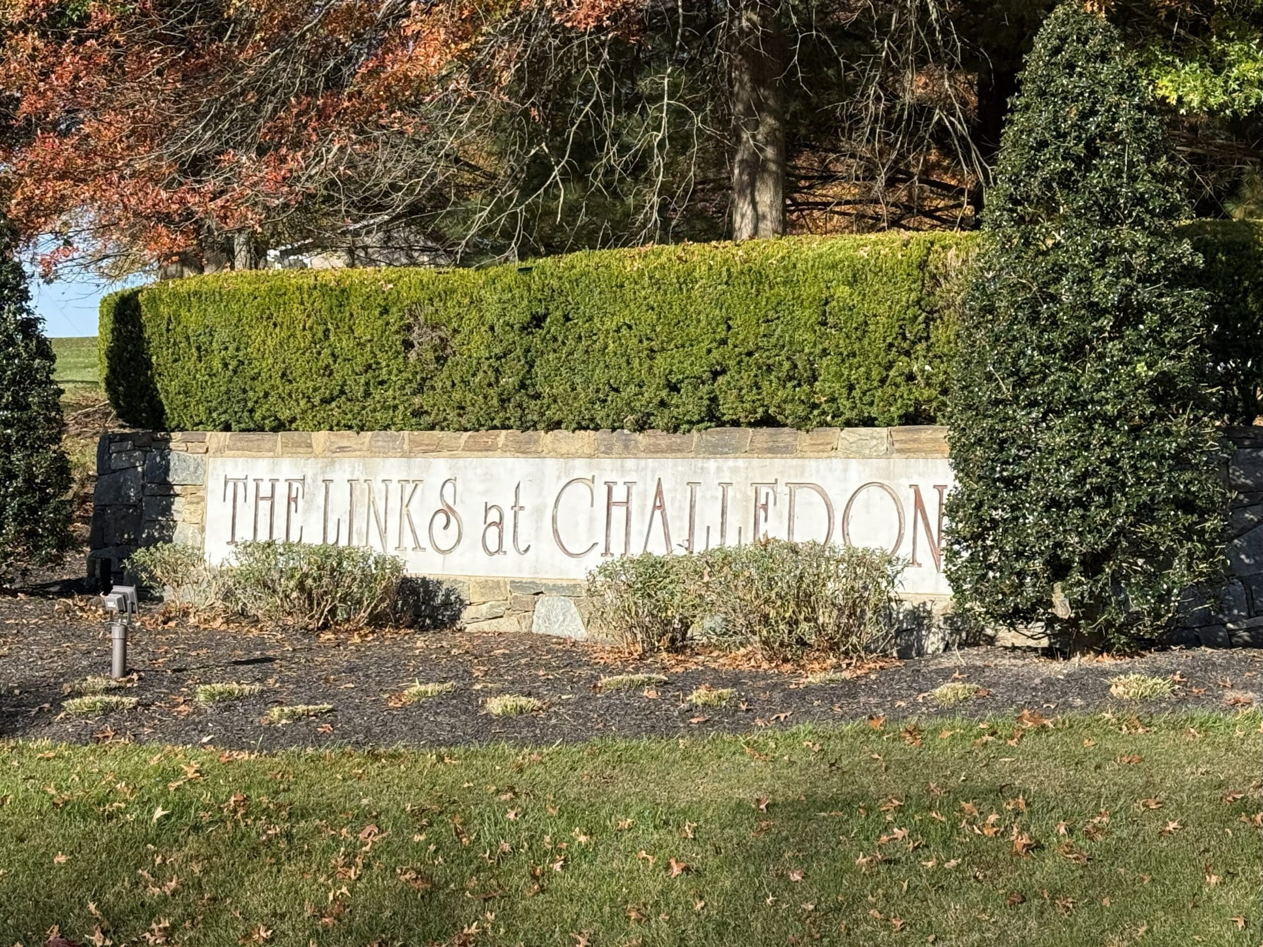 the Links at Challedon Near Frederick Md a Stone Sign with Neatly Trimmed Hedges and Two Tall Bushes Displays the Words the Links at Challedon Autumn Leaves and Sunlight Add Warmth to the Scene While a Small Light Fixture Highlights the Links at Challedons Welcoming Entrance