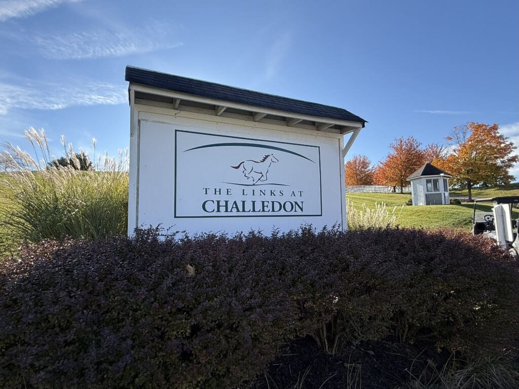 the Links at Challendon Golf Course Review a White Sign with a Horse Logo and Text Reading the Links at Challedon Stands Among Shrubs and Tall Grass in Mt Airy Md in the Background Autumn Trees with Orange Leaves and a Small Shed Sit Under a Bright Blue Sky with Wispy Clouds