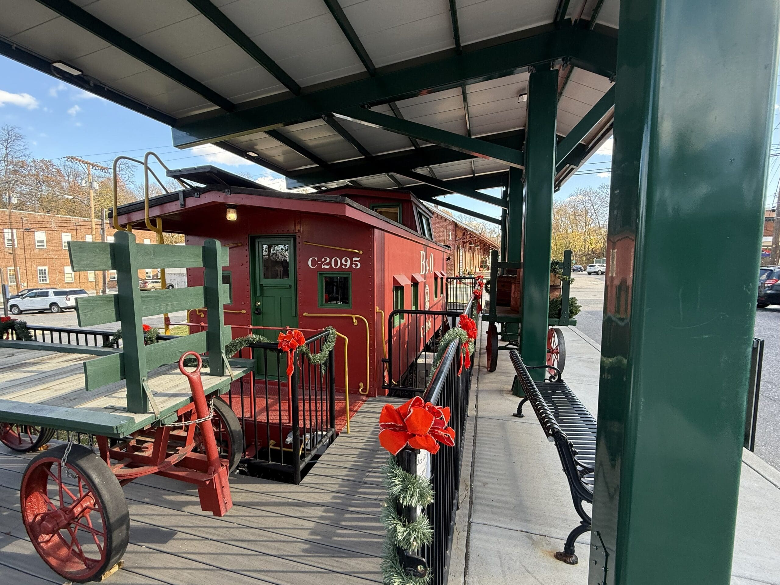 a Red Caboose Train Car Labeled C 2095 Sits on Display Under a Green Metal Canopy in Mt Airy Md Decorative Garlands with Red Bows Adorn Black Railings While a Wooden Luggage Cart Rests Nearby Buildings and Benches Are Visible in the Background
