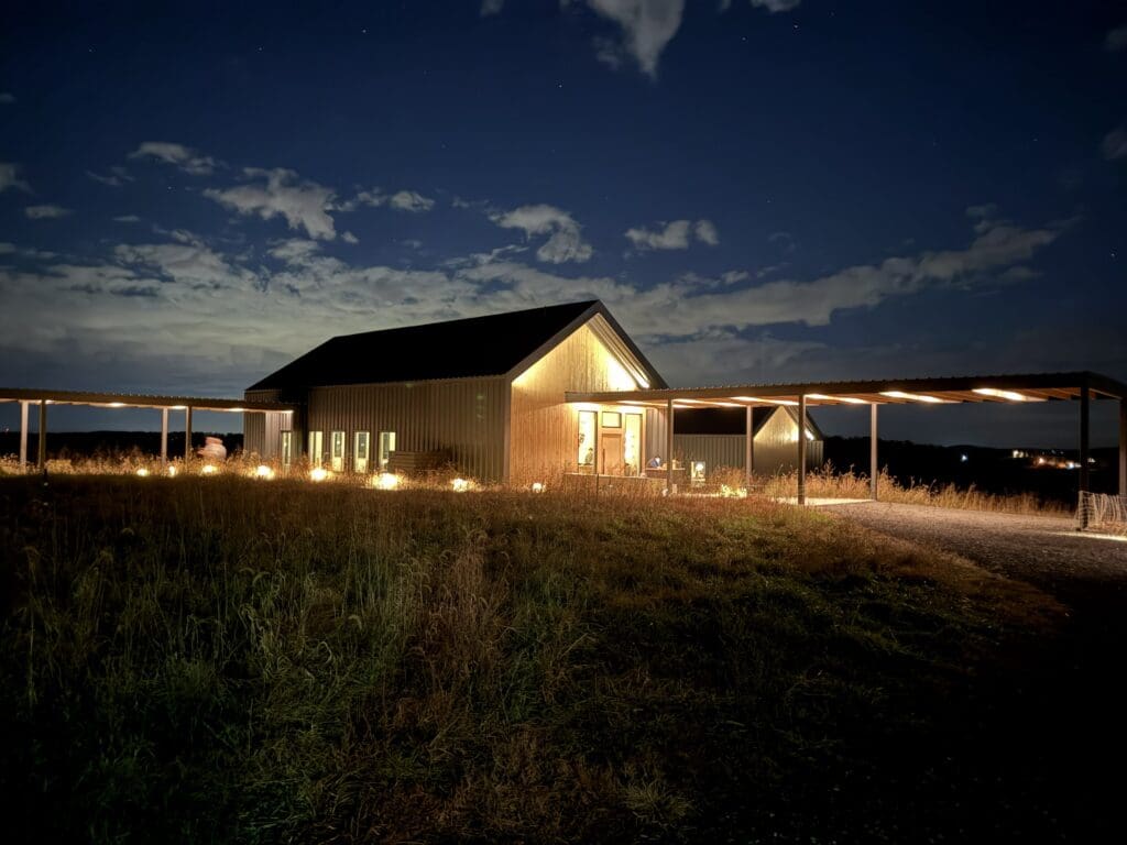 Burnt Hill Farm a Warmly Lit Barn Style Building Stands Alone at Night Under a Partly Cloudy Sky in Mt Airy Md Soft Lights Illuminate Windows and a Covered Walkway While Tall Grass Surrounds the Structure Creating a Peaceful Rural Scene