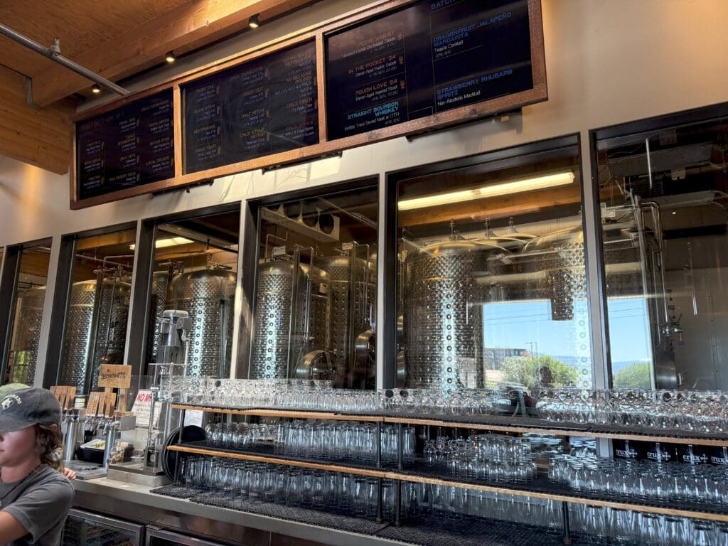 a Modern Brewery Bar at Crux Fermentation Project with Shelves of Clean Glassware Shiny Brewing Tanks Visible Through Windows and Digital Menu Boards Above a Worker in a Cap Stands at the Left As Natural Light Streams In Illuminating Bar and Tanks