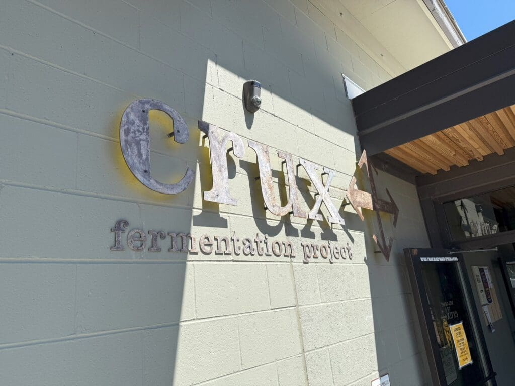 a Metal Crux Fermentation Project Sign is Mounted on a Pale Green Wall Outside a Building Sunlight Casts Shadows Across the Wall and Sign Part of a Wooden Awning and the Entrance Door is Visible on the Right Side