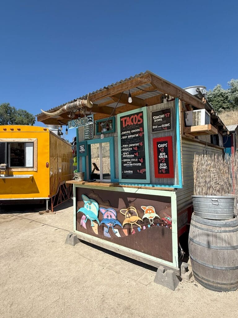 a Small Taco Stand with a Menu Board Displaying Taco Options Chips and Drinks Sits Beside a Bright Yellow Crux Fermentation Project Food Truck the Stand Features Colorful Cartoon Faces a Rustic Roof and a Wooden Barrel Under the Clear Blue Sky