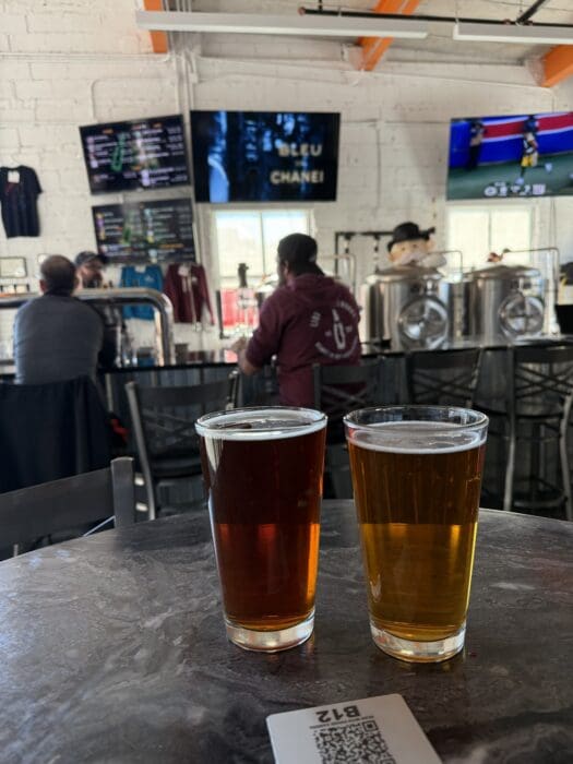 Two Pint Glasses of Beerone Dark Amber One Light Goldensit on a Round Table in a Mt Airy Md Brewery in the Background People Relax at the Bar with Stainless Steel Brewing Tanks Tv Screens and a Menu Board on a White Brick Wall