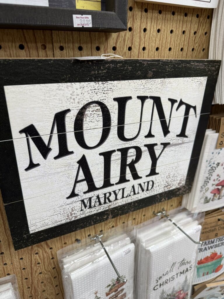 a Wooden Sign with a Distressed White Background and Bold Black Letters Reads Mt Airy Md the Sign Hangs on a Pegboard Wall Above Various White Cloth Items with Different Designs Price Tags Are Visible at the Top