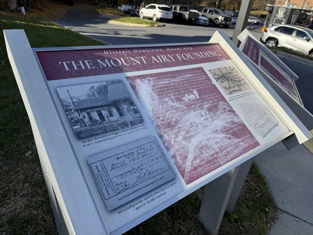 Interpretive Sign Titled the Mount Airy Founding in Historic Downtown Mt Airy Md Features Historical Photos Handwritten Documents and Text Located Outdoors by a Sidewalk the Sign Overlooks Parked Cars and Grass in the Background