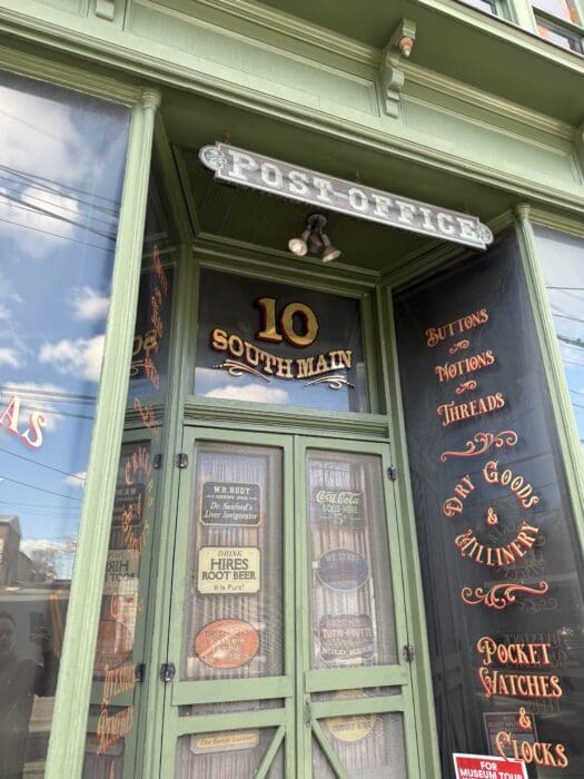 a Vintage Style Post Office Storefront at 10 South Main in Mt Airy Md Featuring Green Trim Glass Doors and Decorative Gold and Red Lettering Advertising Buttons Dry Goods Pocket Watches and Other Old Fashioned Items Clouds Reflect on the Glass