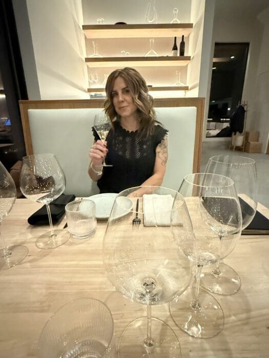 a Woman with Wavy Brown Hair in a Black Dress Sits at a Restaurant Table Holding a Wine Glass from Burnt Hill Farm She is Smiling Slightly the Table Has Several Empty Wine Glasses Plates and Cutlery Shelves with Glassware Are on the Wall Behind Her